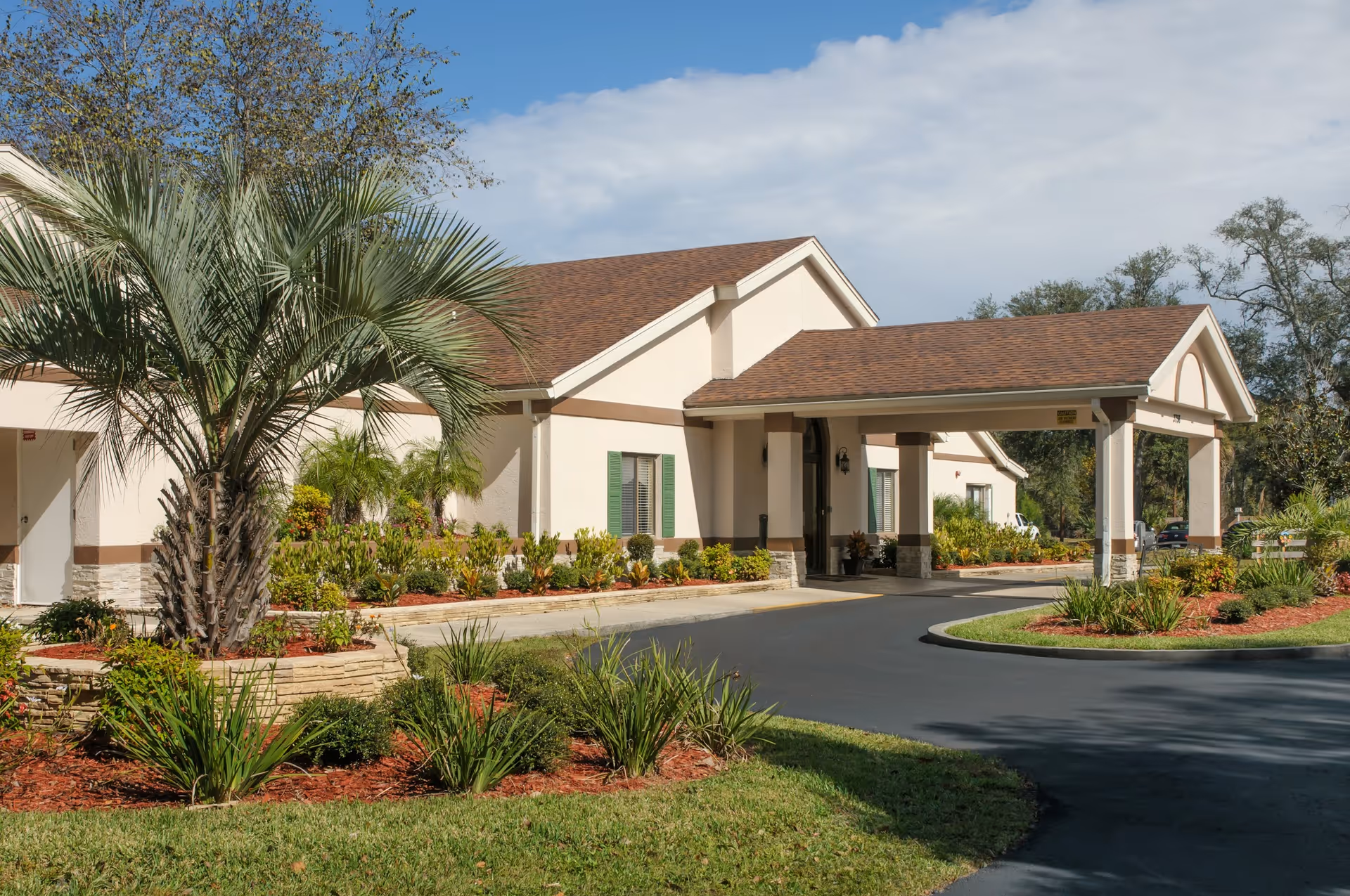 Entrance of a single-story senior living facility with a covered drive-through portico, palm tree and landscaped grounds.