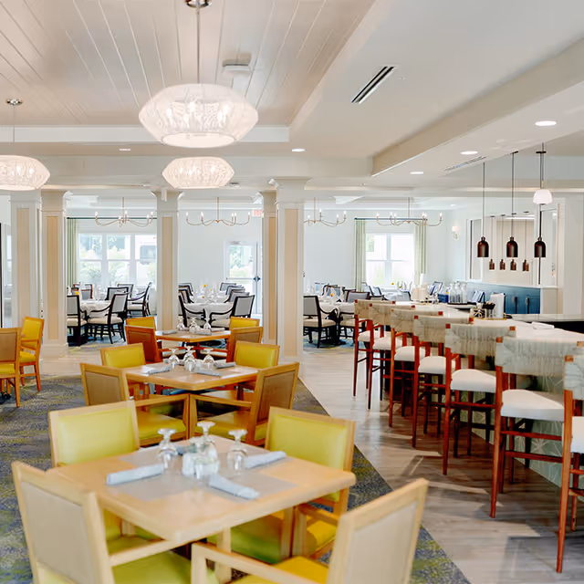 Bright and spacious dining room with multiple tables and chairs arranged neatly. The room features yellow cushioned chairs around wooden tables in the foreground, and a bar area with high stools on the right. Large windows allow natural light to fill the space, and modern pendant lights hang from the ceiling.