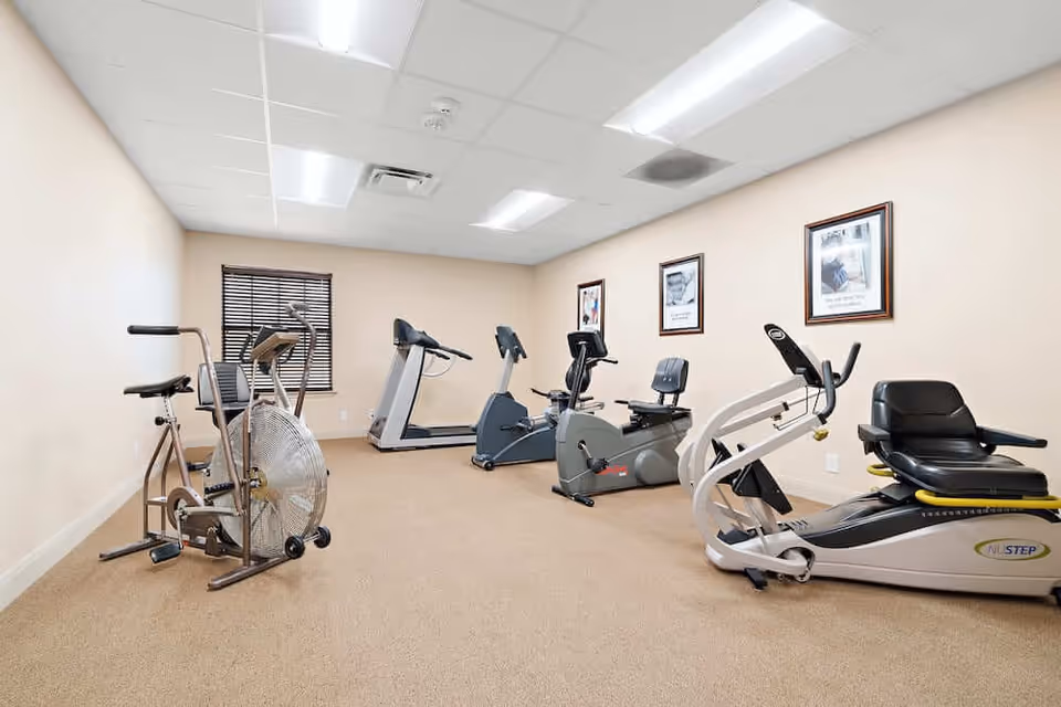 A small fitness room with beige walls and carpeted floor containing several exercise machines including stationary bikes, a treadmill, and a NuStep recumbent cross trainer. The room has a window with blinds and framed pictures on the wall.