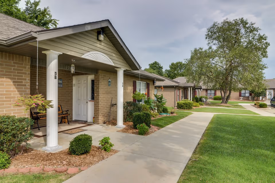 Covered porch and entrance of a single-story brick senior living building with a sidewalk, landscaping, and trees.