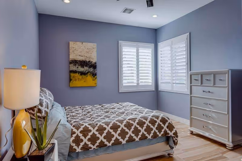 A neatly made bedroom with a patterned bedspread, nightstand lamp and plant, shuttered windows, and a chest of drawers against lavender walls.