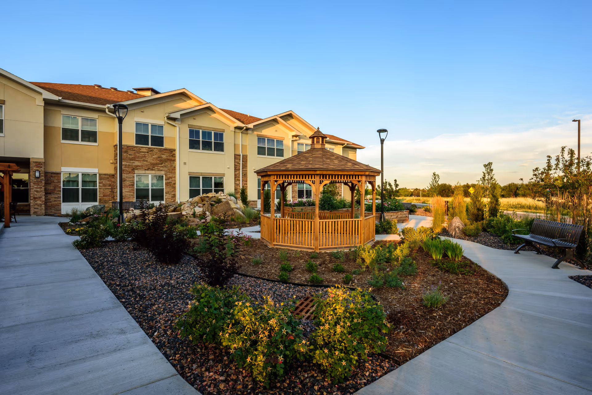 Outdoor garden area of a senior living facility with a wooden gazebo in the center, surrounded by landscaped plants and bushes. There are paved walkways, benches, and a two-story building with beige and stone exterior in the background under a clear blue sky.