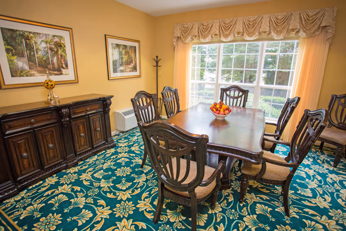 A dining room with a wooden dining table surrounded by six matching chairs with cushioned seats. A bowl of fruit is placed in the center of the table. The room has a large window with sheer curtains and a decorative valance, allowing natural light to brighten the space. The floor is covered with a patterned carpet in teal and gold tones. Against one wall is a dark wooden sideboard with decorative items on top, and two framed pictures hang above it.