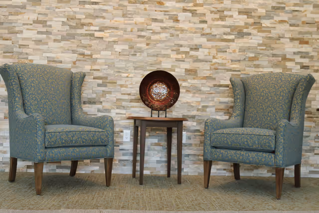 Two upholstered wingback chairs flank a small wooden side table with a decorative plate set against a stacked-stone accent wall.