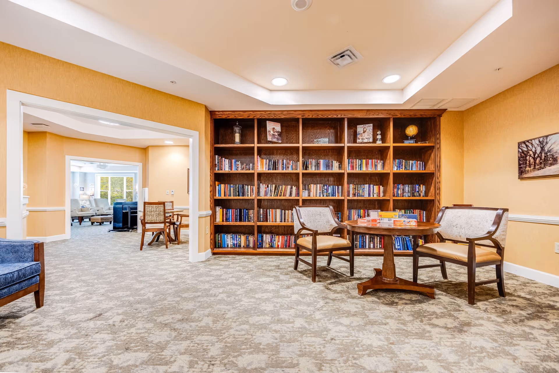 A bright communal library-style lounge with a tall wooden bookshelf, a round table with chairs and board games, and carpeted floors.