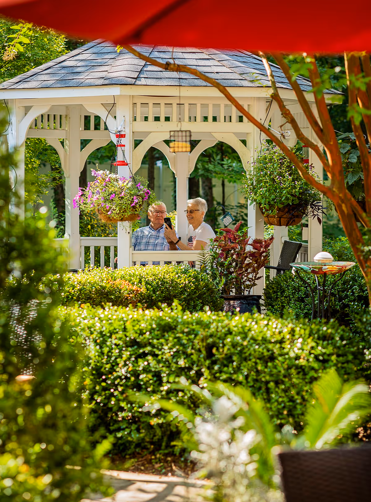 Two elderly people sitting and smiling inside a white gazebo surrounded by lush green bushes and hanging flower baskets in a garden setting.