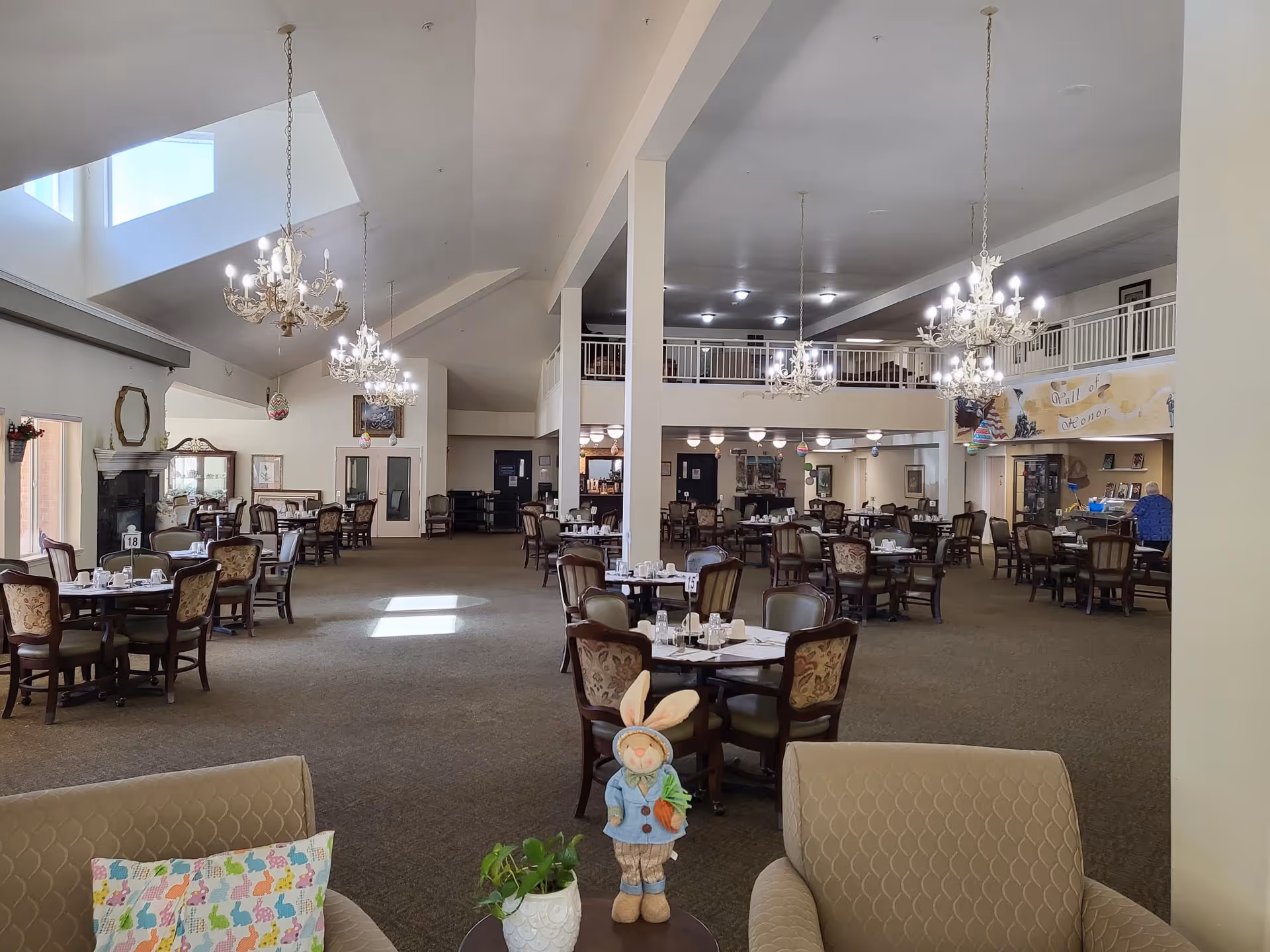 Spacious dining room with multiple round tables and chairs arranged neatly. The room features high ceilings with several chandeliers hanging down, large windows allowing natural light, and a cozy seating area with armchairs and a small table with a decorative bunny and a plant. There is a mezzanine level with railings and a 'Wall of Honor' display on the far wall.