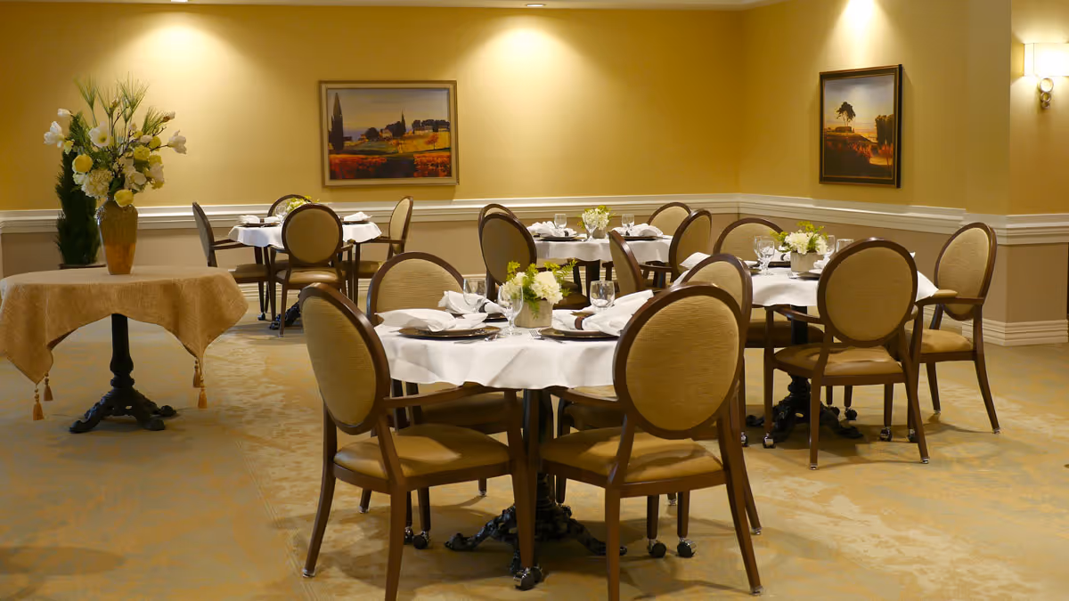 A dining room with round tables covered in white tablecloths, each set with plates, napkins, glasses, and small floral centerpieces. The room has beige walls with two framed landscape paintings and a small table with a vase of flowers in the corner.