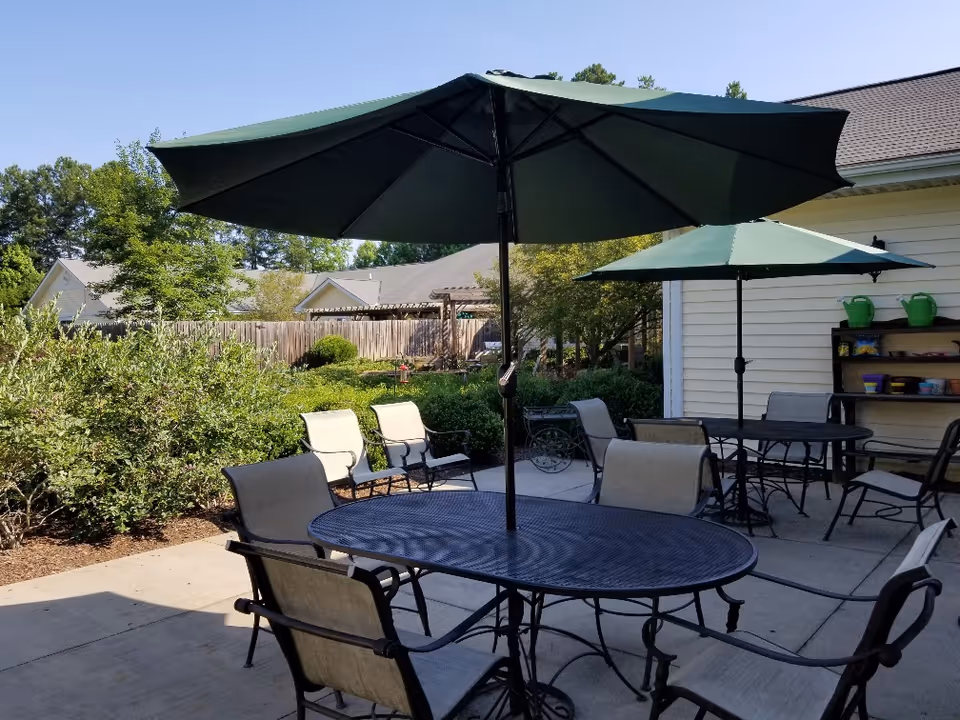 Outdoor patio with round metal tables, chairs, and large green umbrellas beside a garden and building.