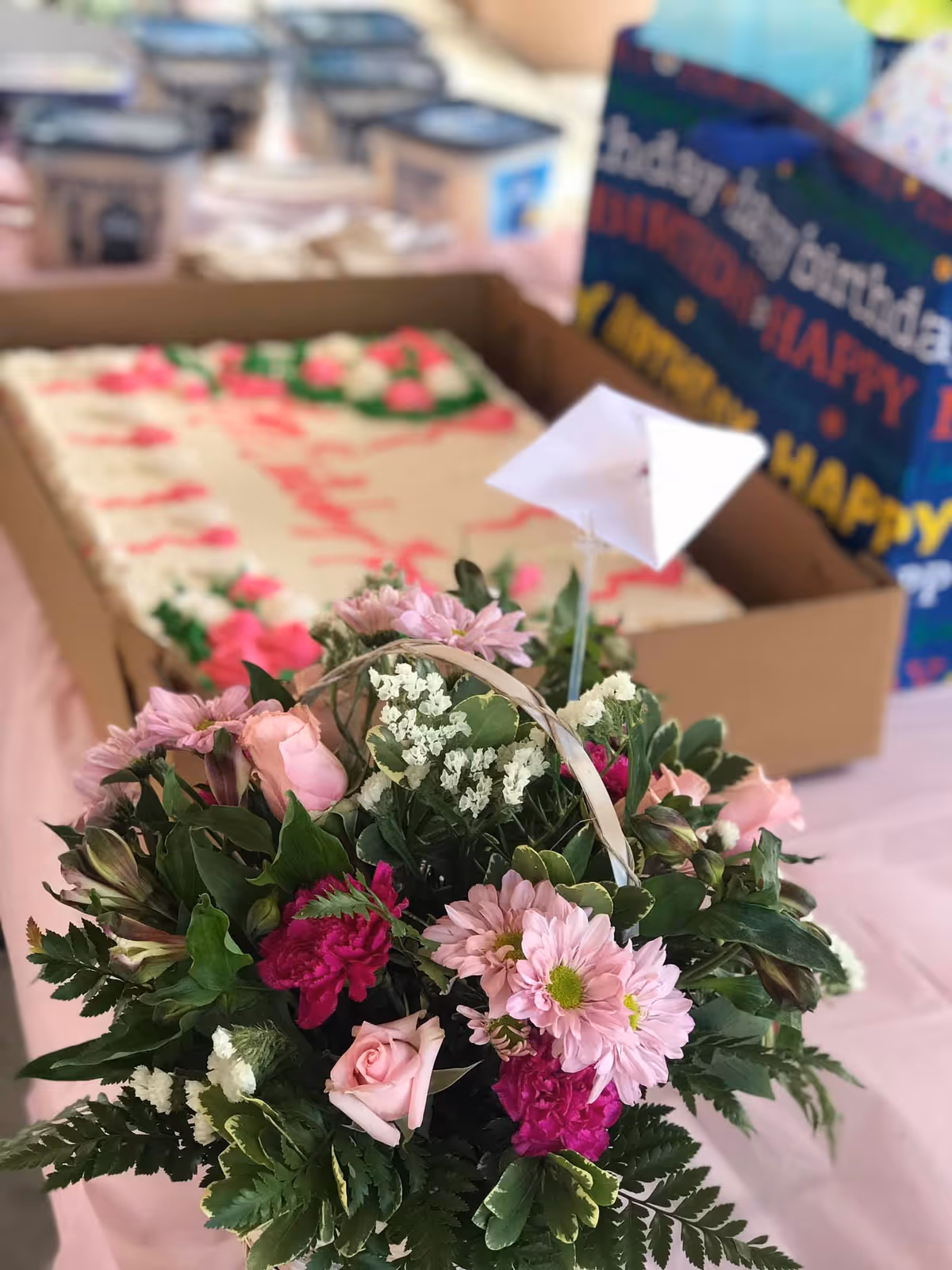 A close-up of a floral arrangement with pink roses, purple flowers, white filler flowers, and green leaves in the foreground. In the background, there is a rectangular cake with white frosting and pink decorations, along with a colorful gift bag with birthday-themed text.