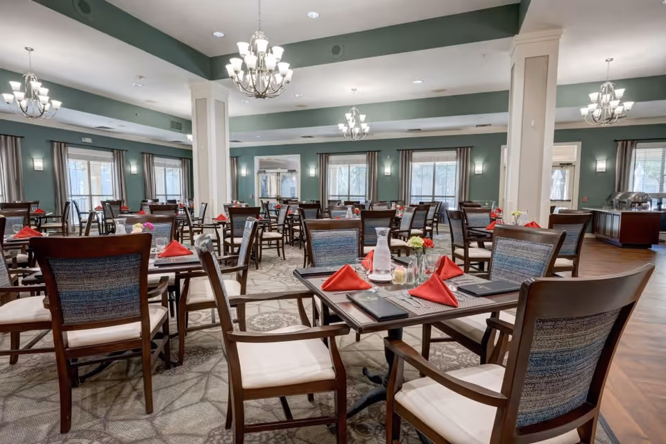 A spacious dining room with multiple wooden tables and chairs arranged neatly. Each table is set with red folded napkins, placemats, menus, and small flower vases. The room has green walls, large windows with curtains, and elegant chandeliers hanging from the ceiling providing warm lighting.