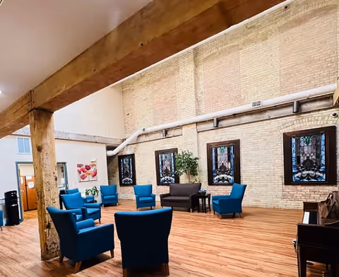 A spacious common area with wooden floors and exposed brick walls featuring stained glass windows. The room contains several blue upholstered armchairs arranged in a circle, a dark brown loveseat, a small table with a plant, and a piano on the right side. Large wooden beams support the ceiling, and there is a door and a water cooler visible in the background.