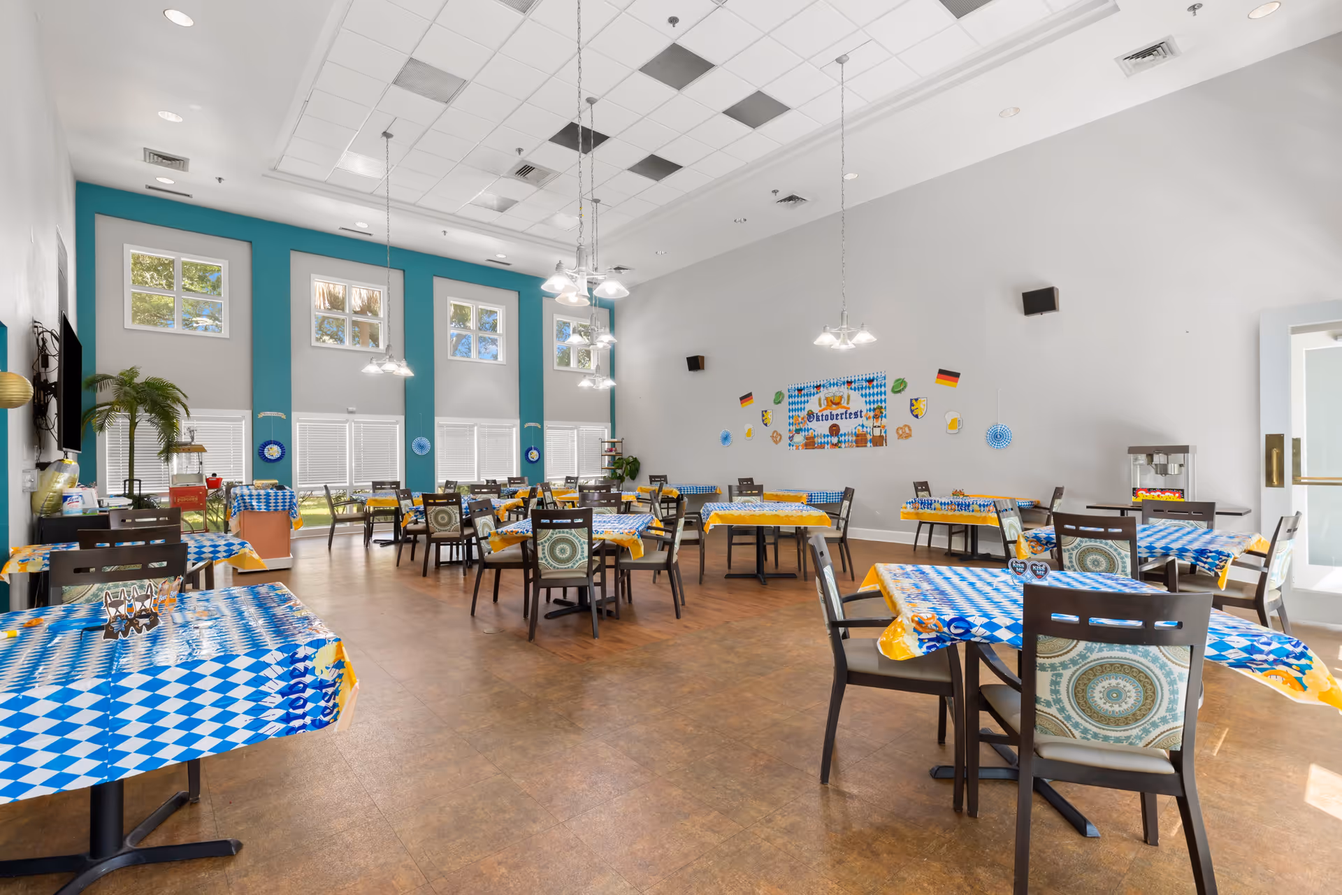 A spacious dining room decorated for Oktoberfest with tables covered in blue and white checkered tablecloths with yellow borders. The room has high ceilings with hanging light fixtures, large windows with white blinds, and walls decorated with Oktoberfest-themed banners and German flags. There are multiple dark wooden chairs with patterned cushions around the tables, and a popcorn machine is visible near the door.