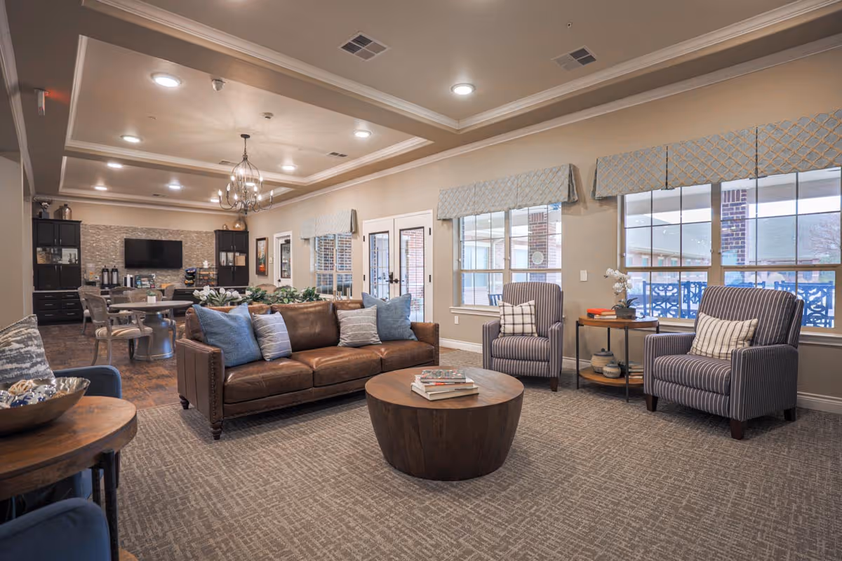 A spacious and well-lit living room in an assisted living facility featuring a brown leather sofa with blue and gray pillows, two striped armchairs with pillows, a round wooden coffee table with books, and large windows with patterned valances. In the background, there is a dining area with a round table and chairs, a wall-mounted TV, and cabinetry with a coffee station.