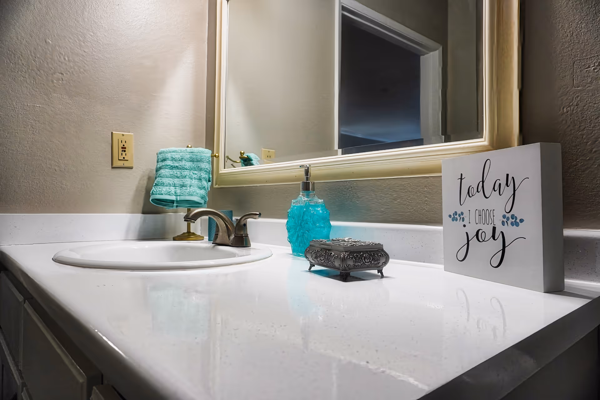 A bathroom countertop with a white sink, silver faucet, turquoise hand towel on a gold holder, turquoise soap dispenser, decorative silver box, and a sign that reads 'today I choose joy' in front of a large mirror.