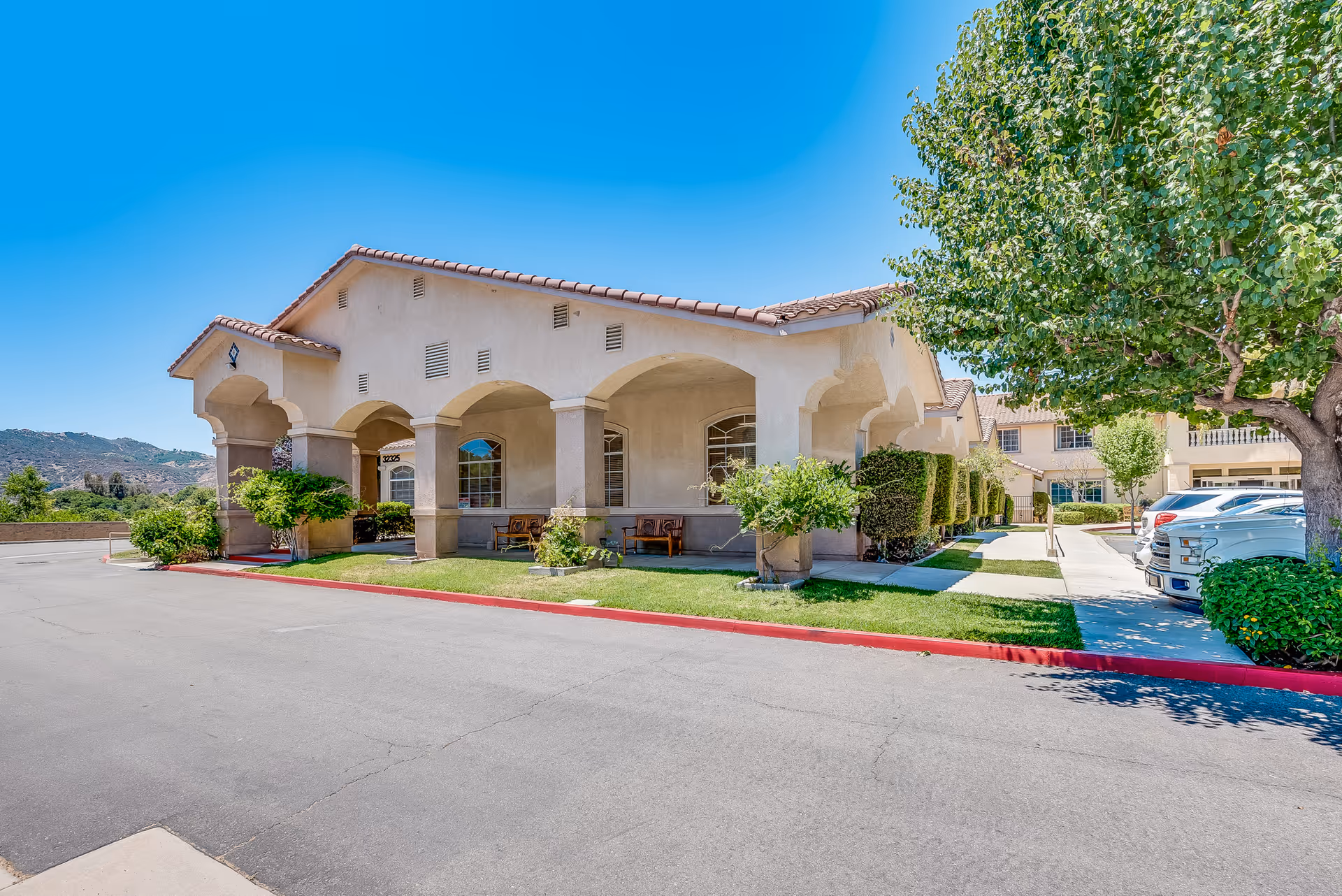 Front exterior of a stucco senior living facility with arched covered walkway, landscaped greenery and parked cars under a clear blue sky.