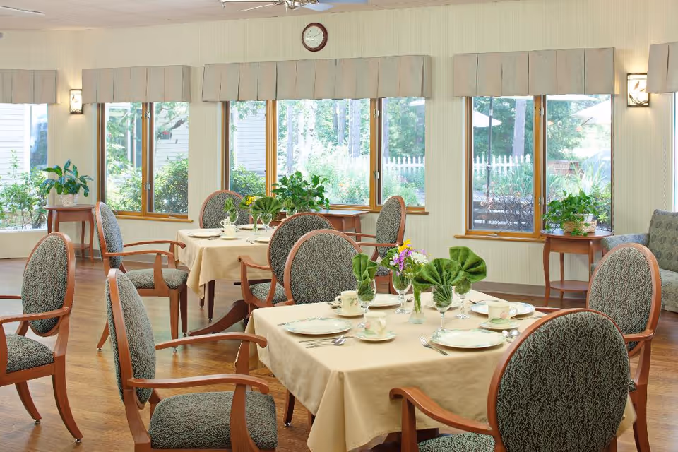 A bright dining room with multiple tables covered in beige tablecloths, each set with plates, cups, silverware, and green folded napkins. The room has large windows with beige valances, wooden framed chairs with patterned upholstery, wooden floors, and several potted plants on small tables near the windows. A clock is mounted on the wall above the windows.