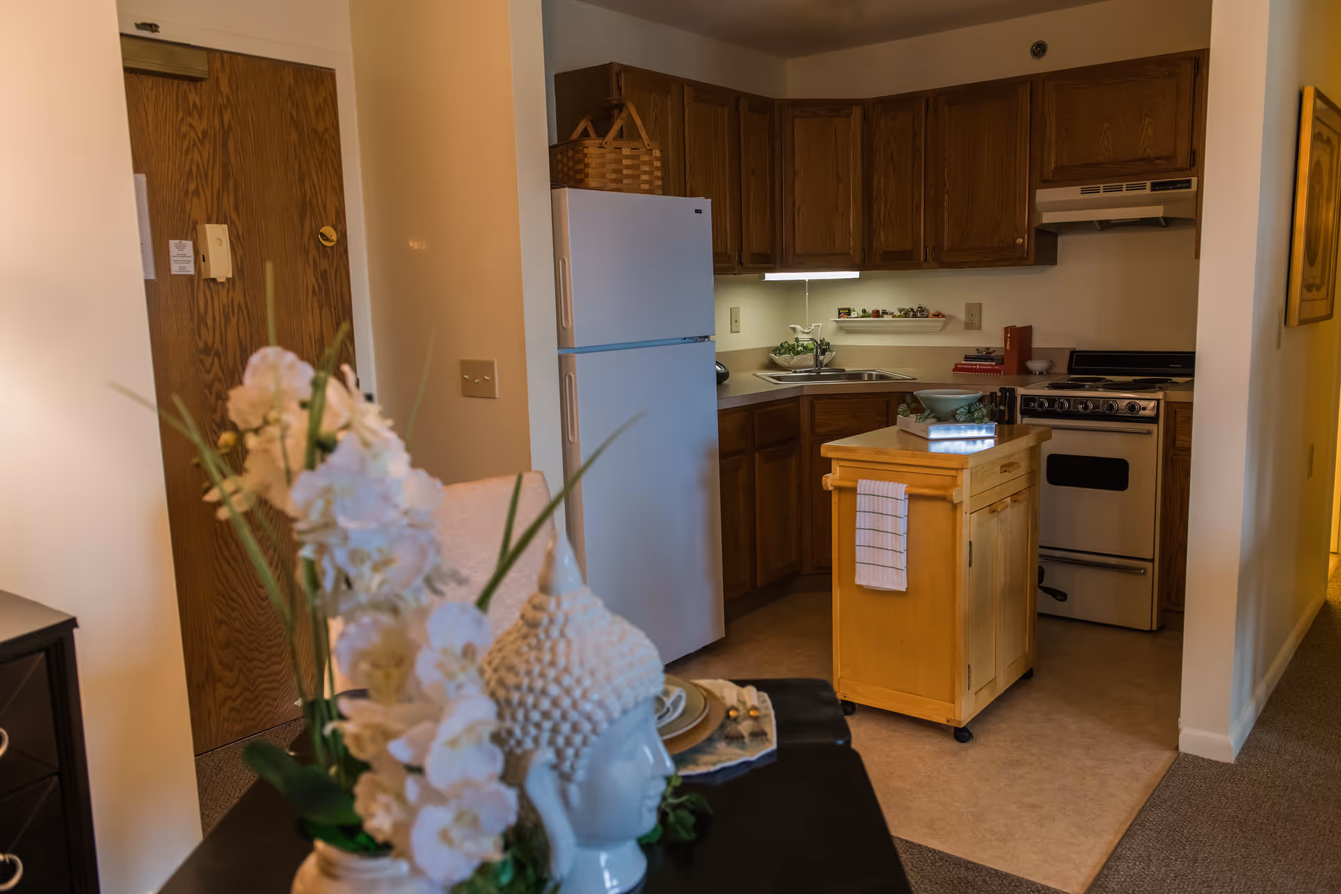 Small kitchenette with wooden cabinets, a white refrigerator, a rolling island cart, and a floral arrangement in the foreground.