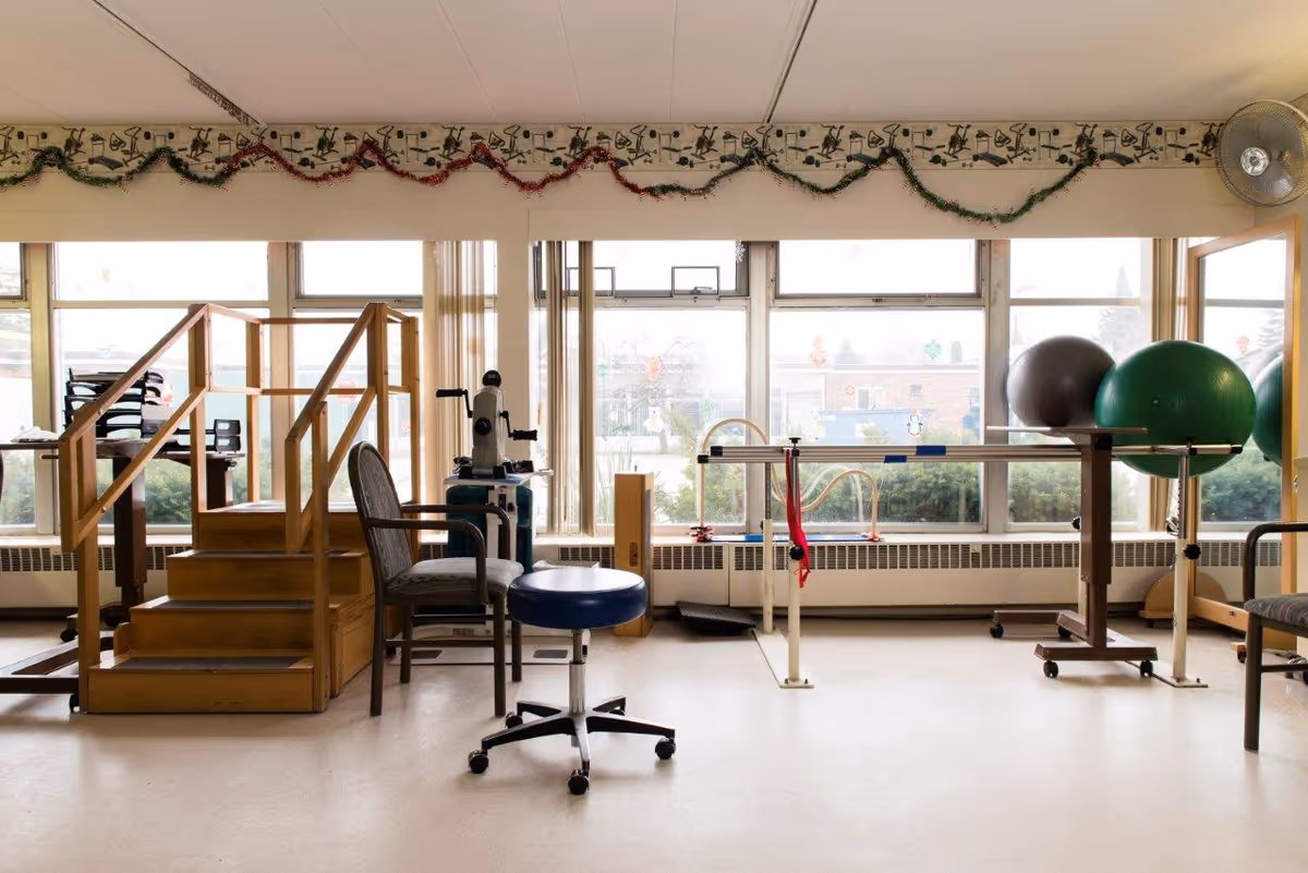 A physical therapy room with wooden stairs for rehabilitation, chairs, exercise balls, parallel bars, and large windows decorated with garlands and holiday stickers.