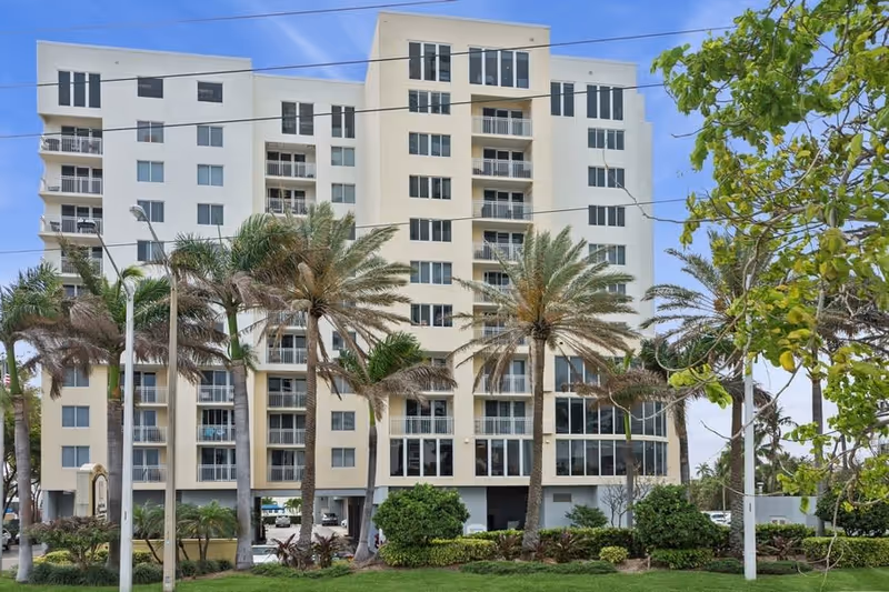 Exterior view of a multi-story residential building with balconies, surrounded by palm trees and greenery under a blue sky.