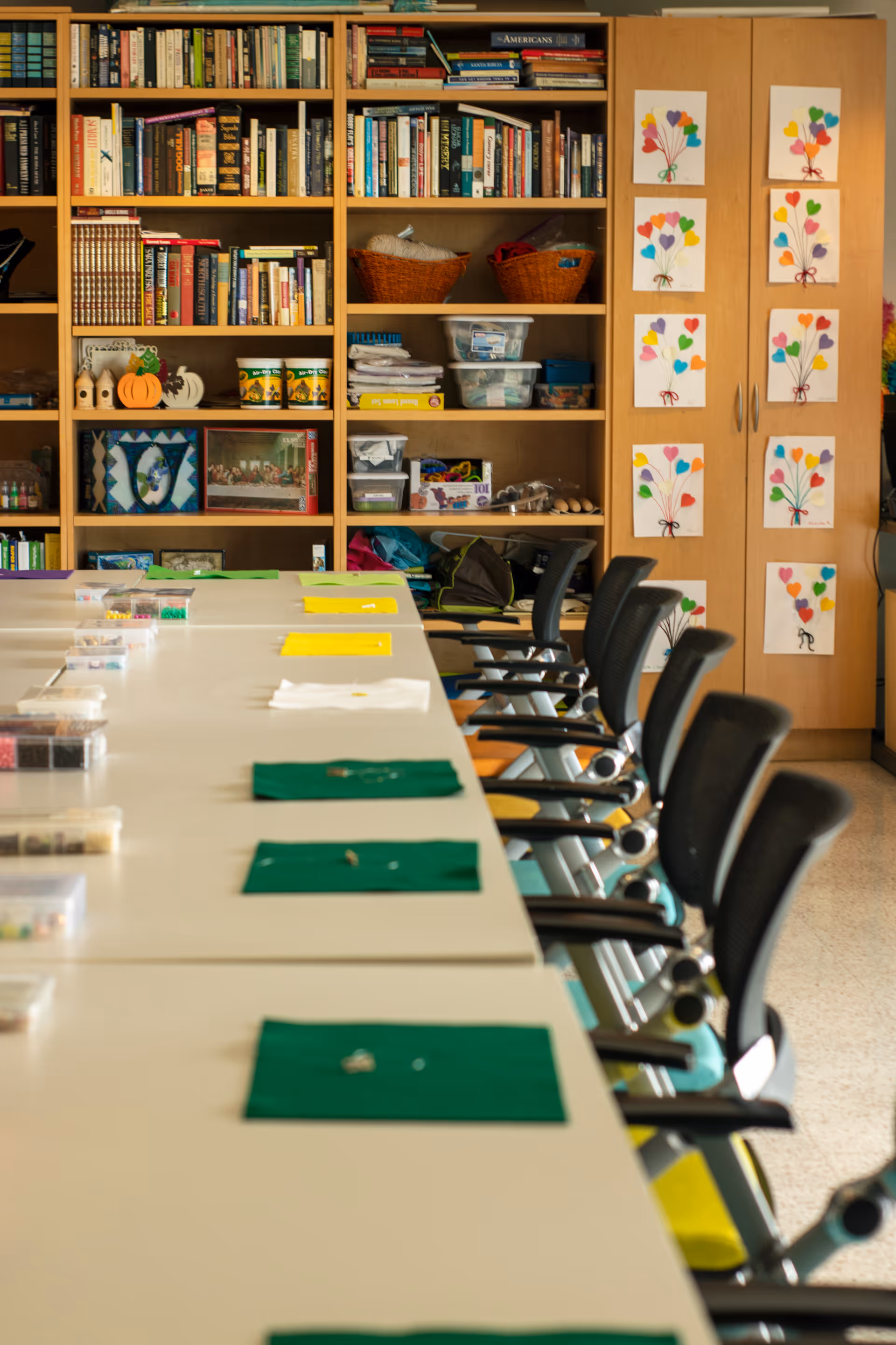 Long table set for crafts with chairs along one side, in a room with bookshelves and cabinets decorated with colorful artwork.