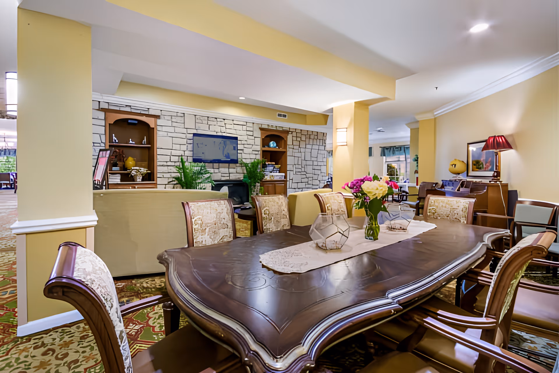 Well-appointed communal dining and lounge area with a large wooden dining table, upholstered chairs, floral centerpiece, and seating area by a stone accent wall.