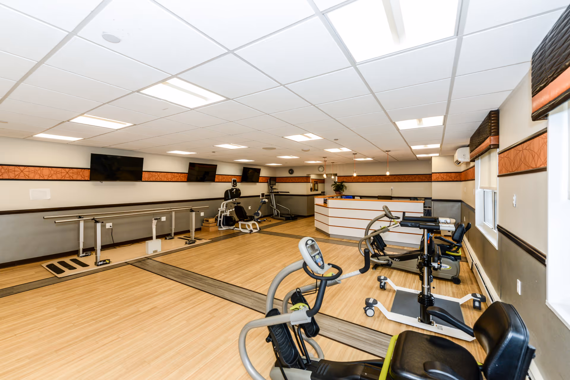 A rehabilitation and exercise room with various exercise equipment including stationary bikes and parallel bars. The room has a light wood floor, white ceiling with fluorescent lights, and several wall-mounted televisions. There is a reception desk at the far end of the room and windows on the right side with blinds partially drawn.
