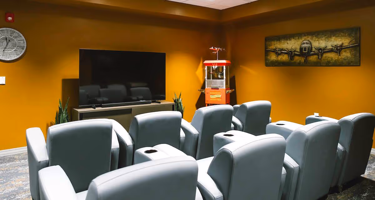 A small theater room with two rows of gray recliner chairs facing a large flat-screen TV mounted on a cabinet. The walls are painted mustard yellow, and there is a popcorn machine in the corner. A decorative airplane painting hangs on the wall, and a round clock is mounted on the left wall.