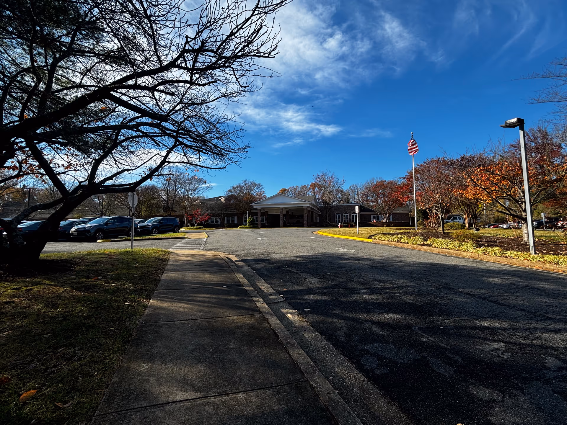 View of the front exterior of Paul Spring Retirement Community building with a driveway, parked cars, leafless trees, and an American flag on a flagpole under a partly cloudy blue sky.
