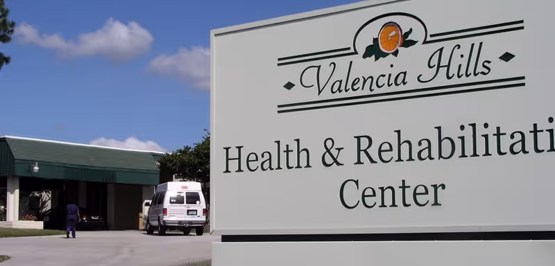Outdoor view of Valencia Hills Health & Rehabilitation Center sign with the facility building and a white van in the background under a blue sky with some clouds.