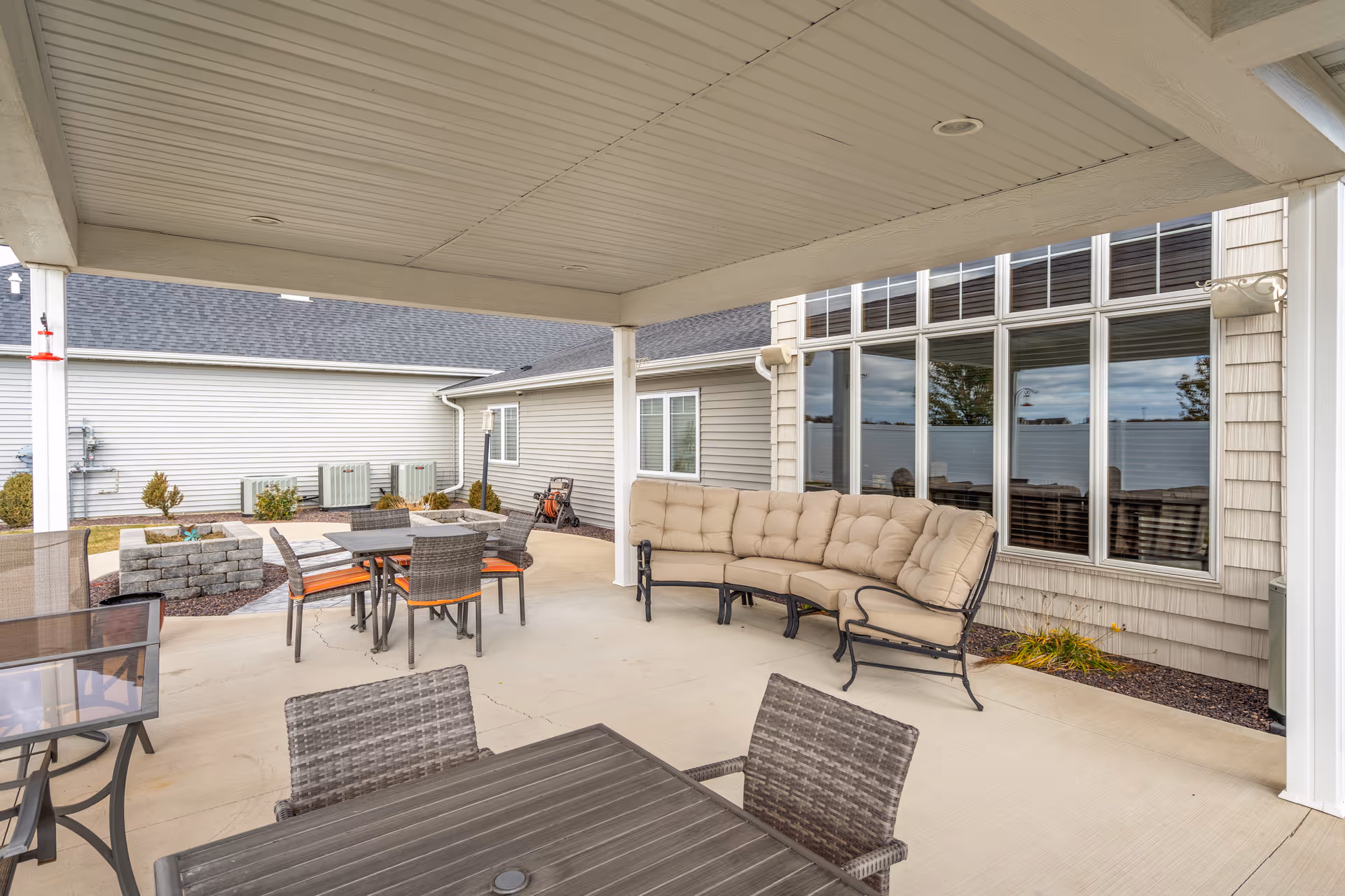 Covered outdoor patio area with cushioned curved sofa, multiple tables with chairs, and a fire pit surrounded by a stone wall. The patio is adjacent to a building with large windows and beige siding.