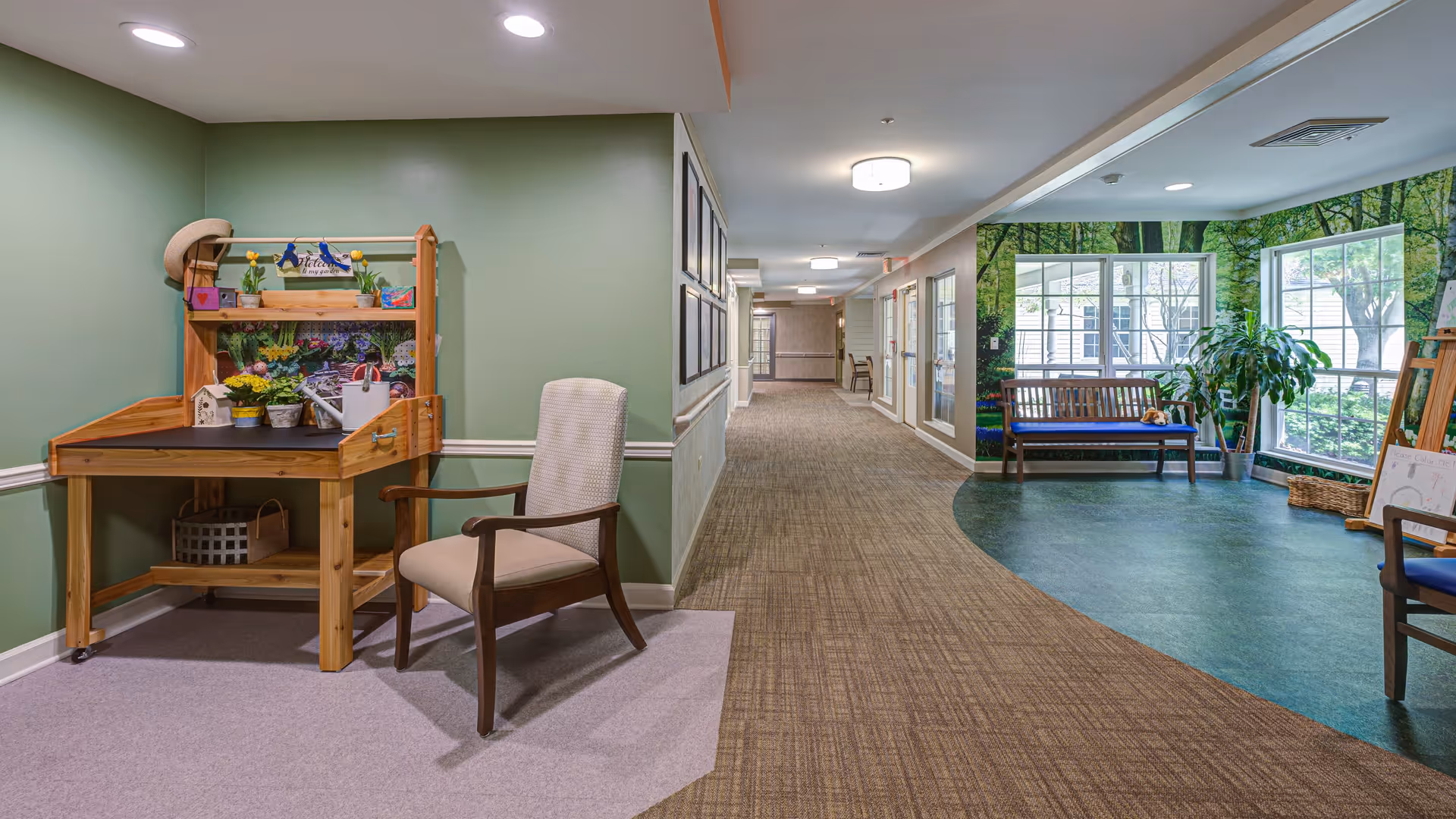 A hallway in a senior living facility with green walls and carpeted floor. On the left side, there is a wooden gardening table with small potted plants and gardening tools, next to a cushioned armchair. On the right side, there is a seating area with a wooden bench and chairs, large windows showing greenery outside, and a mural of trees on the wall.