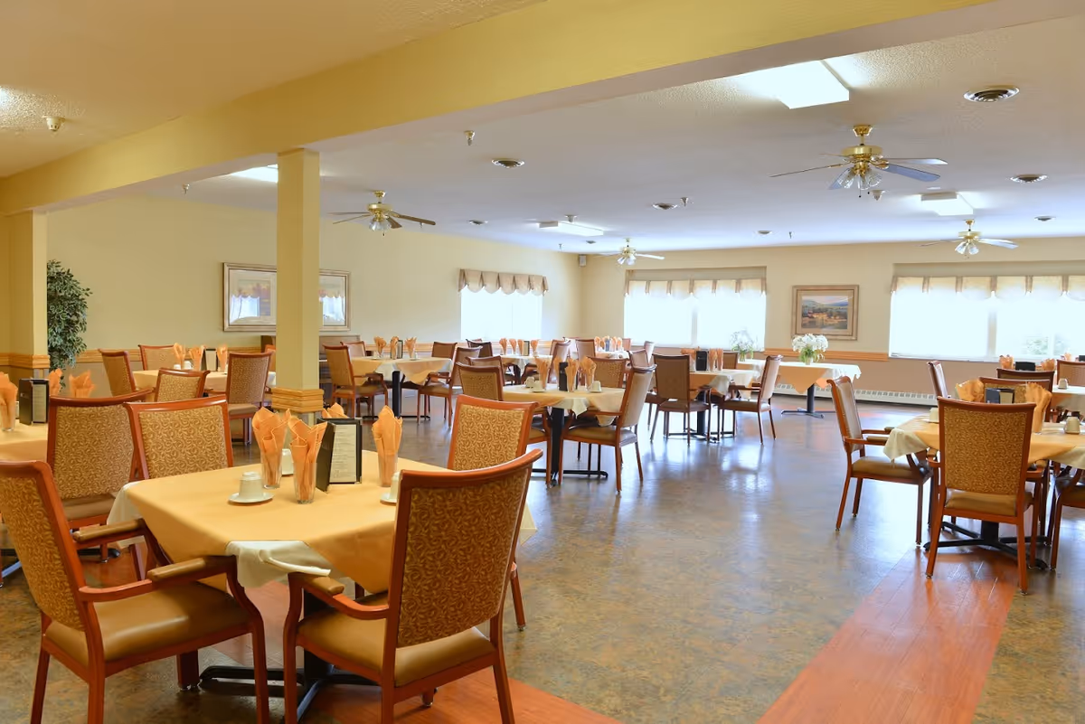 A spacious dining room with multiple tables covered in beige tablecloths and set with folded orange napkins, cups, and menus. The room has large windows with valances allowing natural light to fill the space, ceiling fans, framed artwork on the walls, and comfortable upholstered chairs around each table.