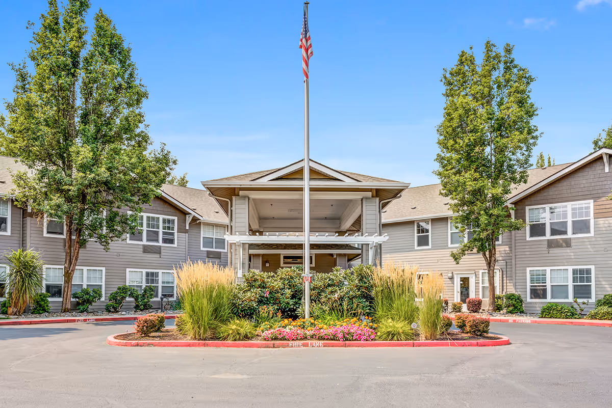 Front exterior view of Woodside Senior Living facility with a covered entrance, an American flag on a flagpole, landscaped greenery, and a clear blue sky.