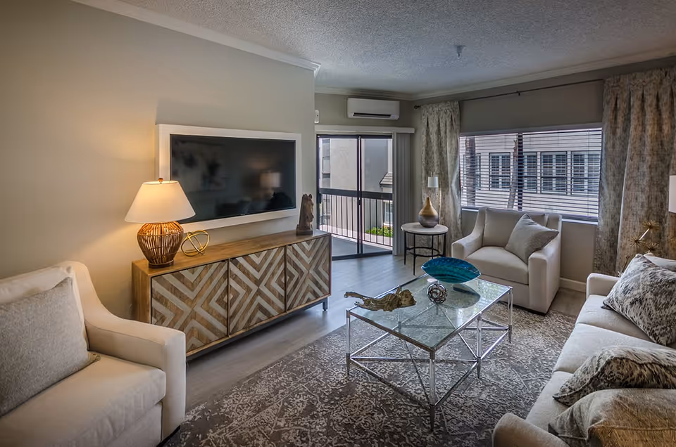 Bright furnished living room with sofa and armchairs, a wall-mounted TV, glass coffee table, and sliding balcony doors.