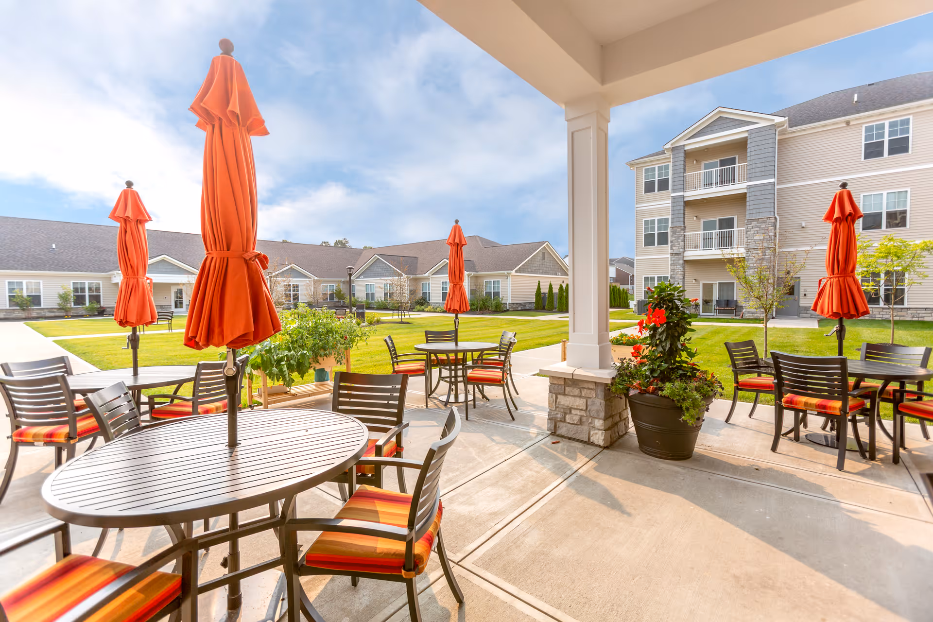 Outdoor patio area with round tables and chairs featuring orange-striped cushions, each table having a closed orange umbrella. The patio overlooks a well-maintained grassy courtyard surrounded by multi-story residential buildings under a partly cloudy sky.
