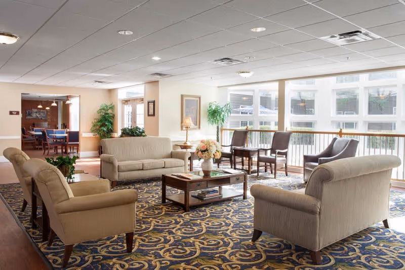 A bright and spacious living room area in a senior living facility with beige upholstered sofas and armchairs arranged around a wooden coffee table with a flower vase. Large windows let in natural light, and there are additional chairs lined up against the window. A patterned carpet covers the floor, and there are plants and framed artwork on the walls.