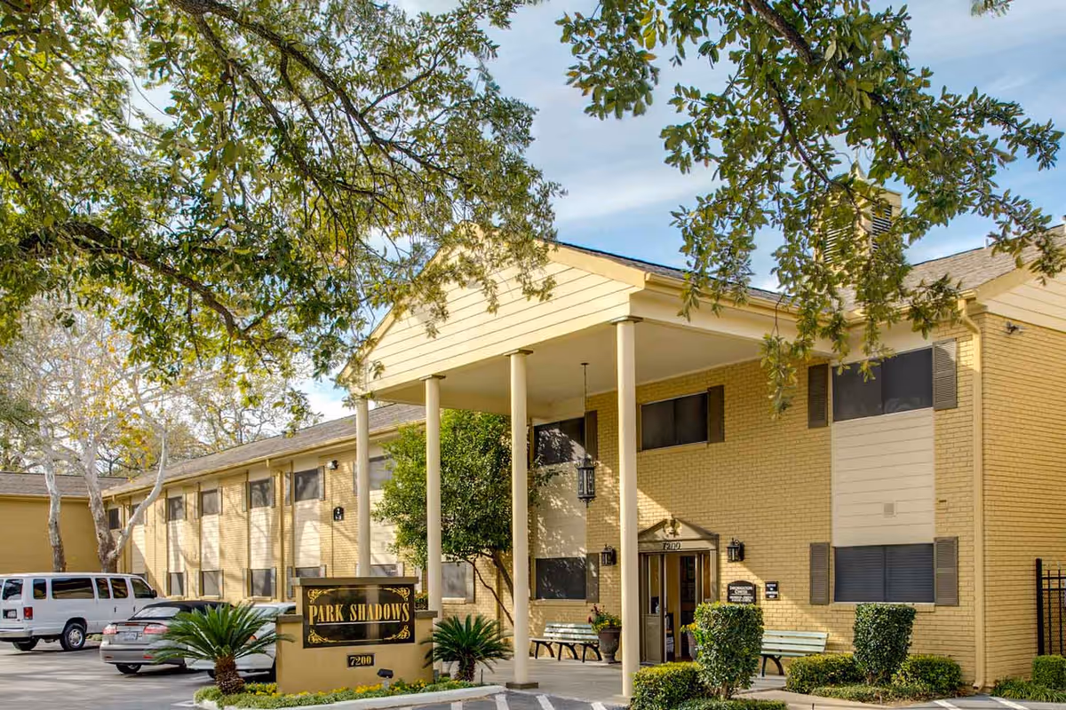Exterior view of Park Shadows Retirement Community building with a covered entrance supported by four columns, surrounded by trees and parked cars in the parking lot.