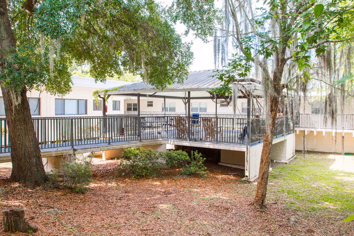 Outdoor patio area with a covered seating space featuring several chairs and tables, surrounded by trees and greenery. The patio is elevated with a railing and is part of a building visible in the background.