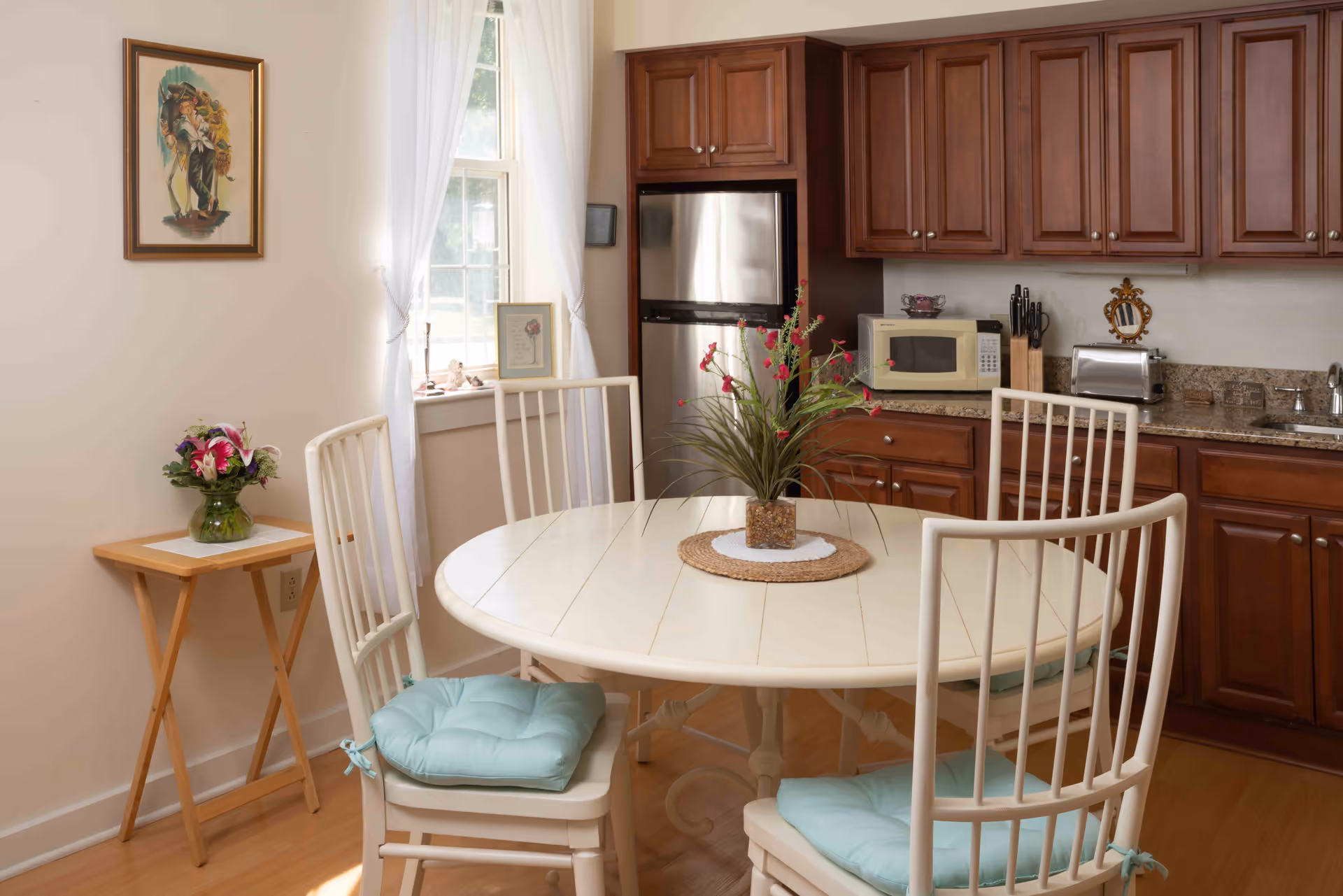 A bright kitchen dining area with a round white table surrounded by four white chairs with light blue cushions. The kitchen features wooden cabinets, a stainless steel refrigerator, a microwave, a toaster, and a sink. A small wooden side table with a vase of flowers is placed near a window with white curtains. A framed picture hangs on the wall above the side table.