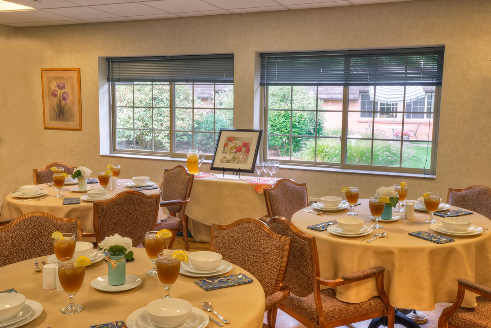 A dining room with round tables covered in beige tablecloths, each set with white bowls, plates, silverware, napkins, and glasses of iced tea garnished with lemon slices. The room has large windows with blinds partially open, showing greenery outside. There are upholstered chairs around the tables and a framed floral picture on a small table near the windows.