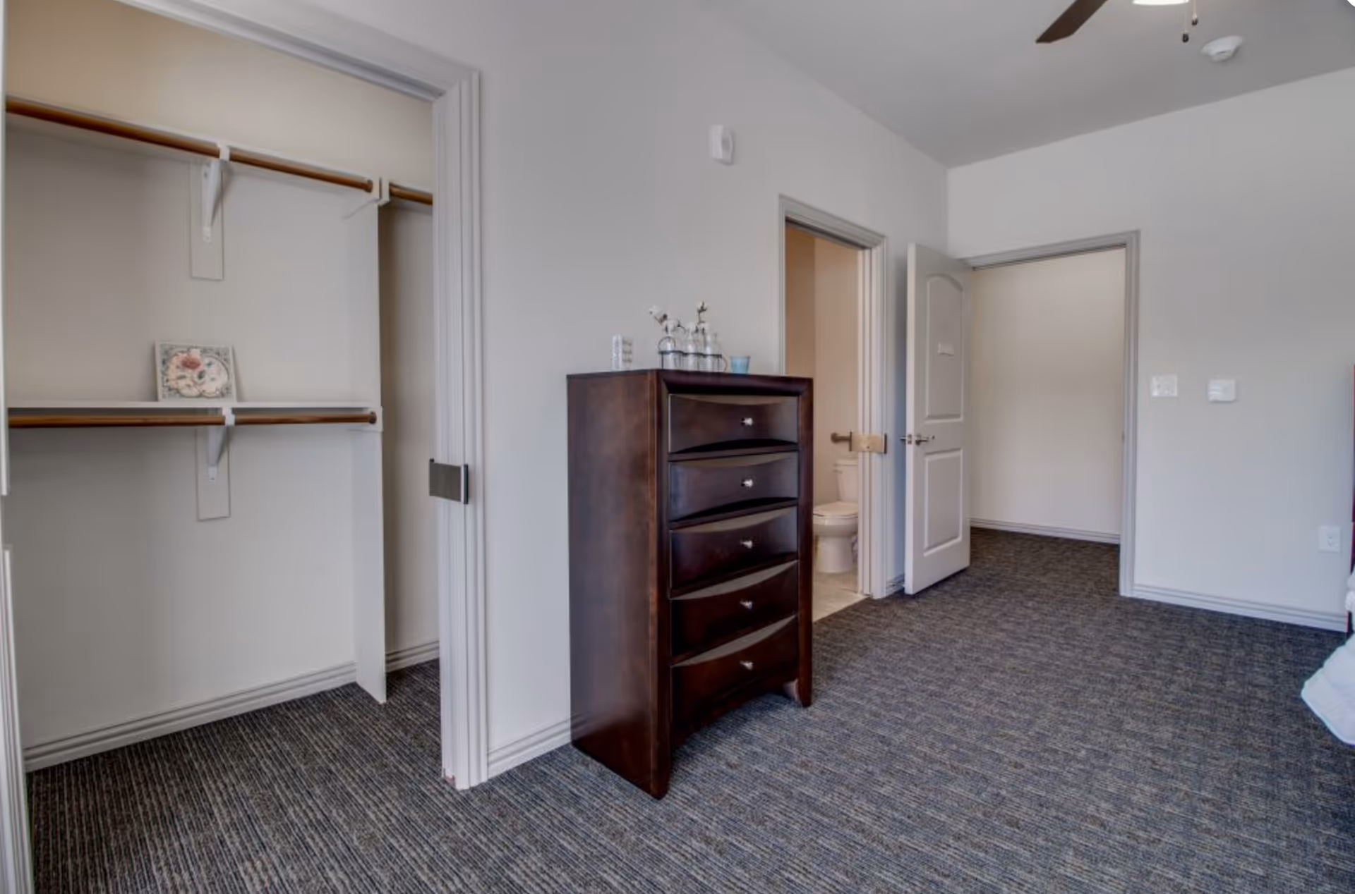 Interior bedroom with an open closet, a tall dark wood dresser, and a doorway leading to a bathroom.