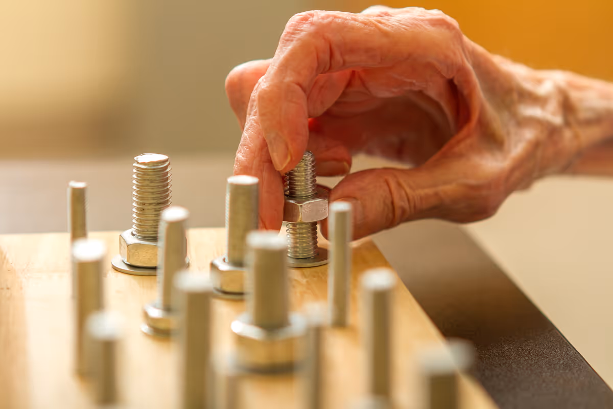 Close-up of an elderly person's hand manipulating a large bolt and nut on a wooden board with multiple bolts and nuts, likely as part of a physical therapy or rehabilitation exercise.