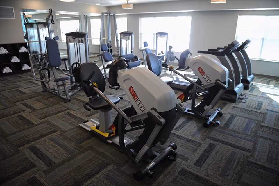 A fitness room with various exercise machines including seated row machines, treadmills, and weight machines. The room has large windows letting in natural light and a shelf with neatly stacked white towels.