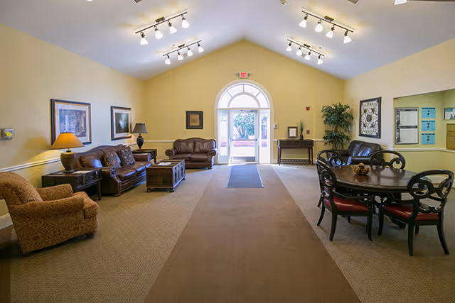 A spacious living room area with beige walls and carpeted floor. The room features multiple seating options including a patterned armchair, two brown leather sofas, and a round wooden table with four chairs. There are framed pictures on the walls, two table lamps, a potted plant, and a large arched glass door at the far end letting in natural light.