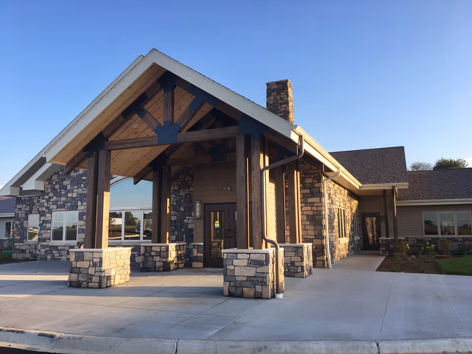 Exterior front entrance of a senior living facility named The Legacy of DeForest, featuring a covered porch with wooden beams and stone pillars, stone and siding exterior walls, and a clear blue sky.