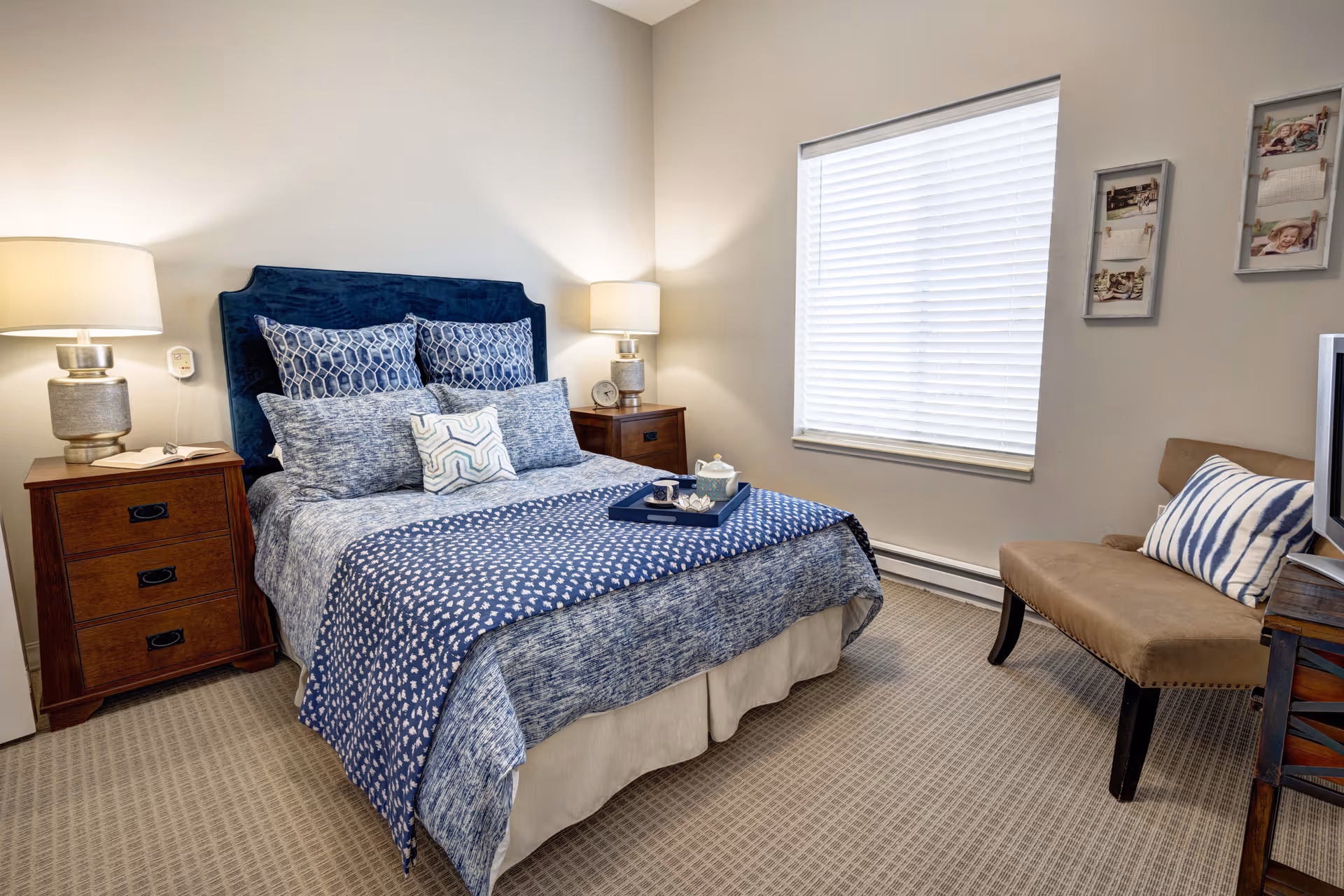 A neatly made bed with blue and white patterned bedding and multiple pillows in a bedroom. There are two wooden nightstands on either side of the bed, each with a lamp. A beige upholstered bench with a striped pillow is placed near a window with closed blinds. The walls are light-colored and decorated with two small picture frames.