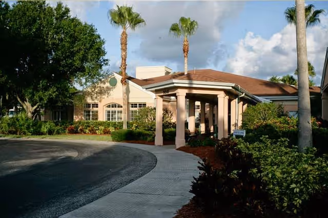 Exterior view of a single-story building with a covered entrance supported by columns, surrounded by palm trees and greenery under a partly cloudy sky.