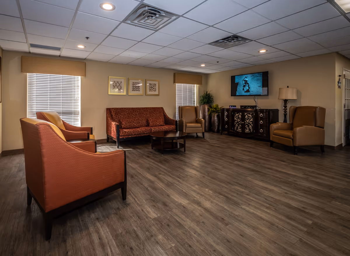 A cozy living room area in a nursing and rehabilitation facility featuring two orange armchairs, a red patterned sofa, two brown armchairs, a round wooden coffee table, a TV mounted on the wall above a decorative wooden cabinet, two windows with blinds and valances, a floor lamp, and potted plants in the corner.