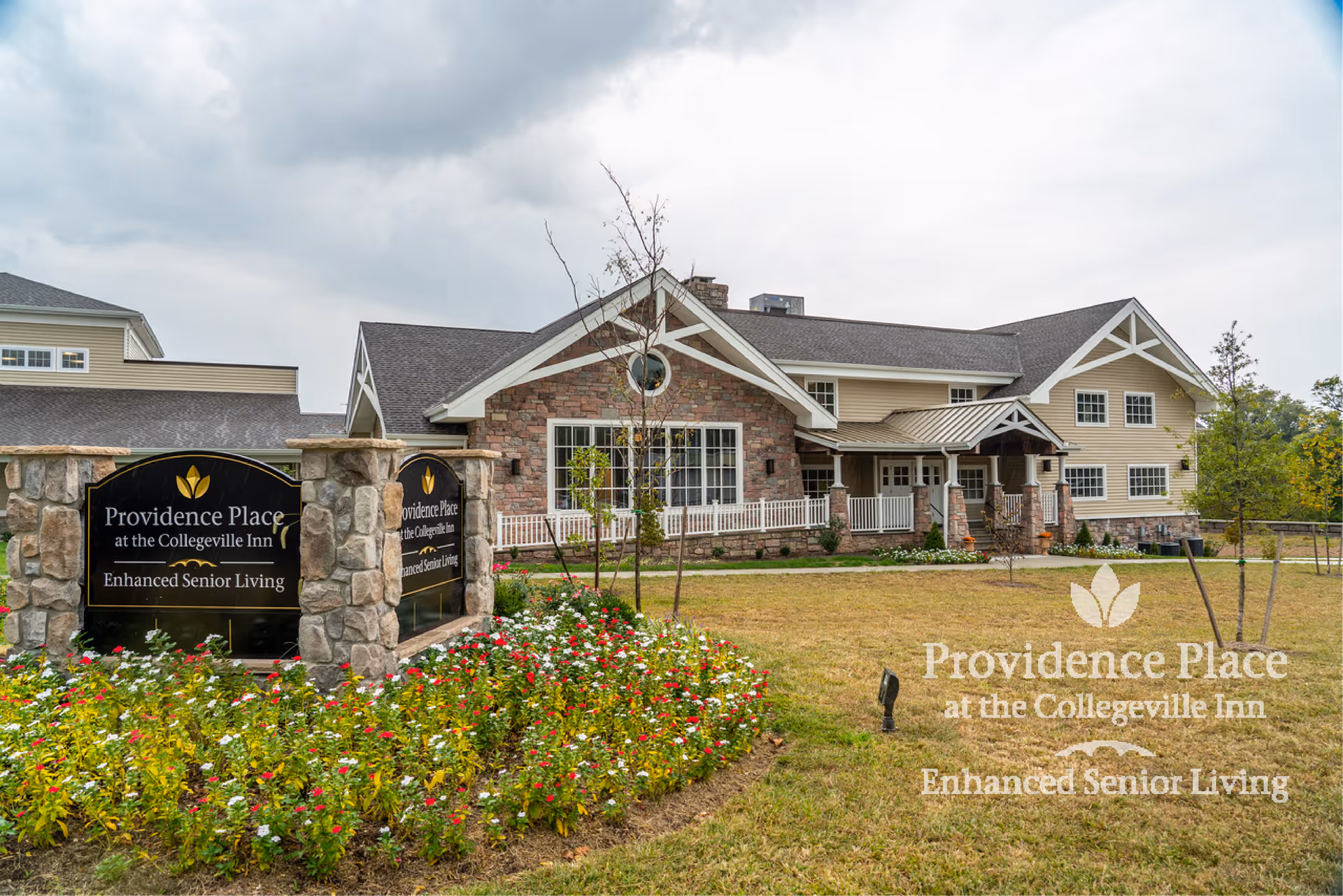 Exterior view of Providence Place Senior Living at the Collegeville Inn, showing a large building with stone and beige siding, a landscaped garden with colorful flowers, and a stone sign with the facility's name and 'Enhanced Senior Living' text.